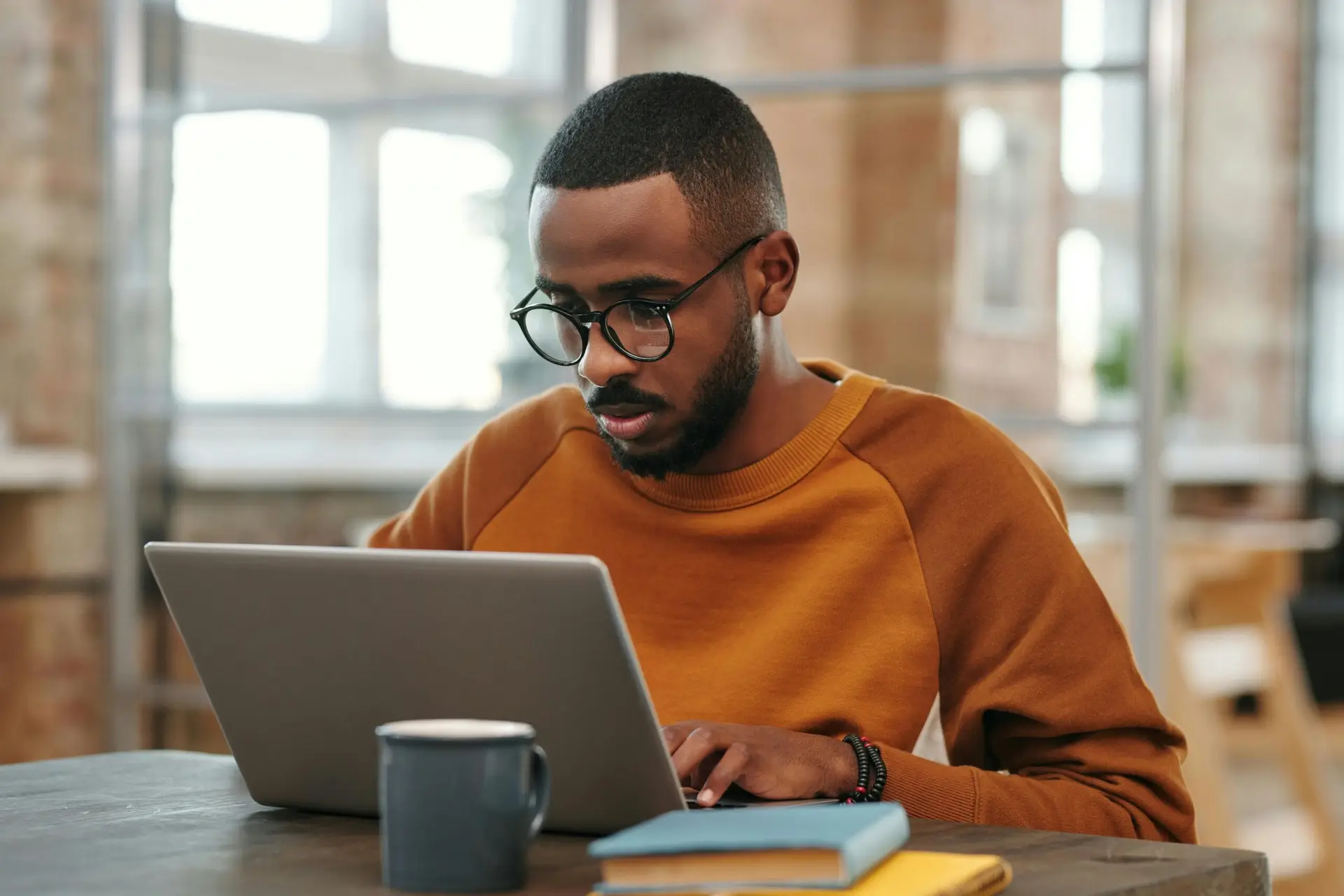 Young man with glasses works on laptop at a wooden table indoors. Warm, productive atmosphere.